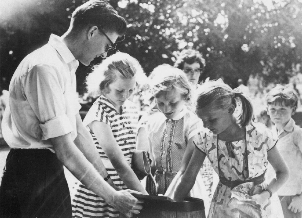 Monochrome photograph of a student helping local children play a game at the 1960 Cripps Hall Summer Fete in aid of the World Refugee Year Appeal.